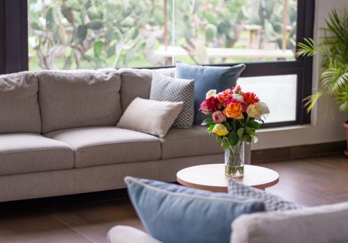 Cozy living room interior providing comfort and relaxation with a gray sofa, throw pillows, and a vibrant flower bouquet on a round coffee table, overlooking a green outdoor garden